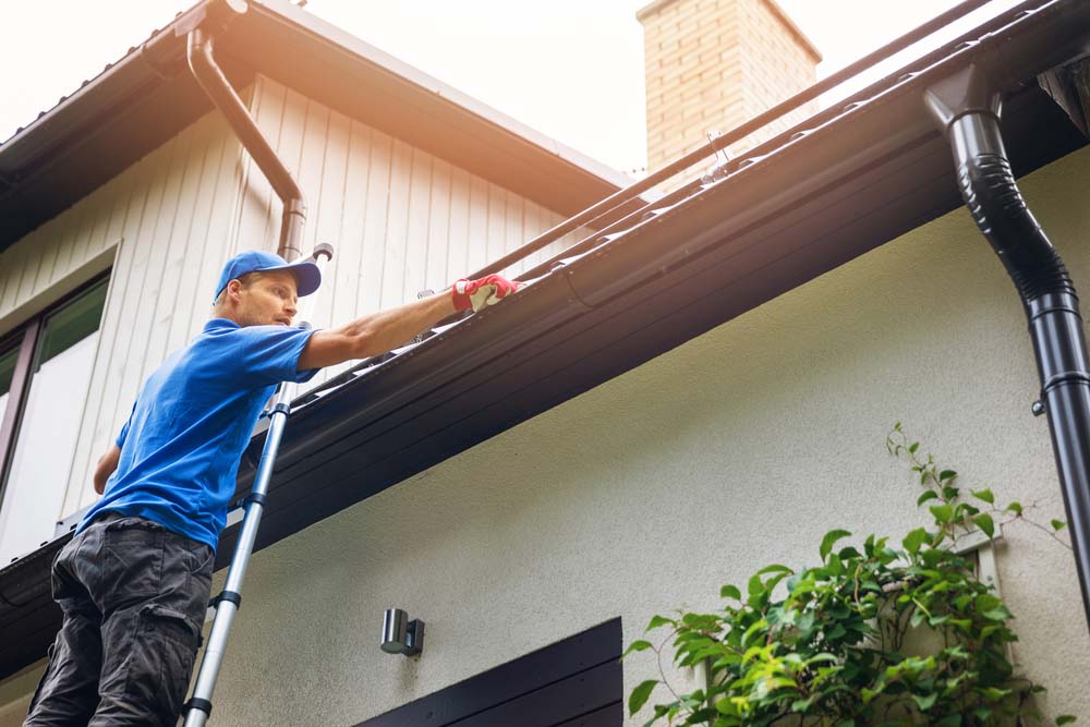 A man in a blue shirt and hat stands on a ladder and cleans the gutters of a house.
