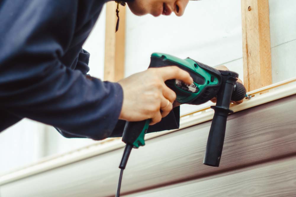 A person installs beige vinyl siding with a power tool.