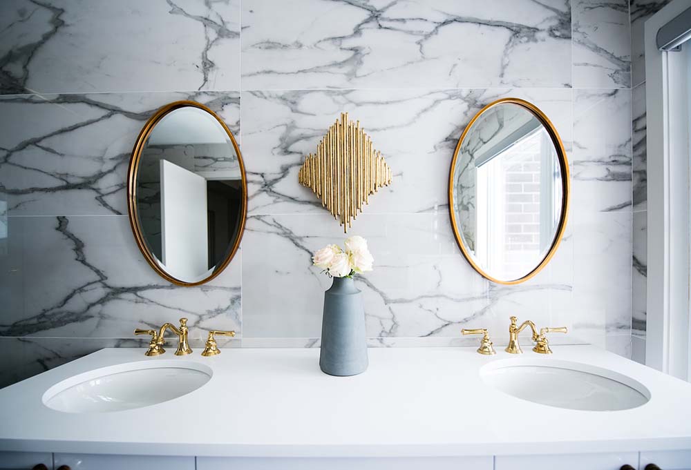 A vanity with two sinks with white countertops against a wall with a marble pattern and two ovular mirrors.